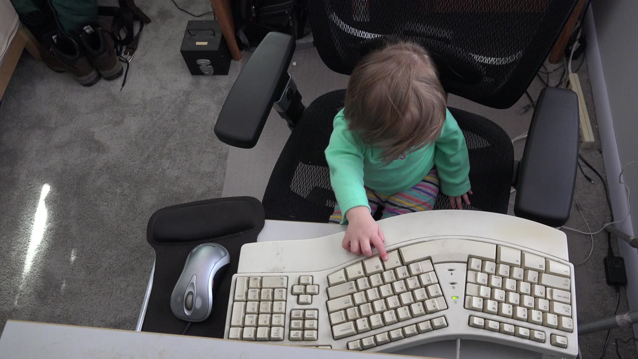 At 0:01 in Westminster, Colorado, the child leans forward in the black mesh chair with one finger resting on the central keys of the split keyboard.
