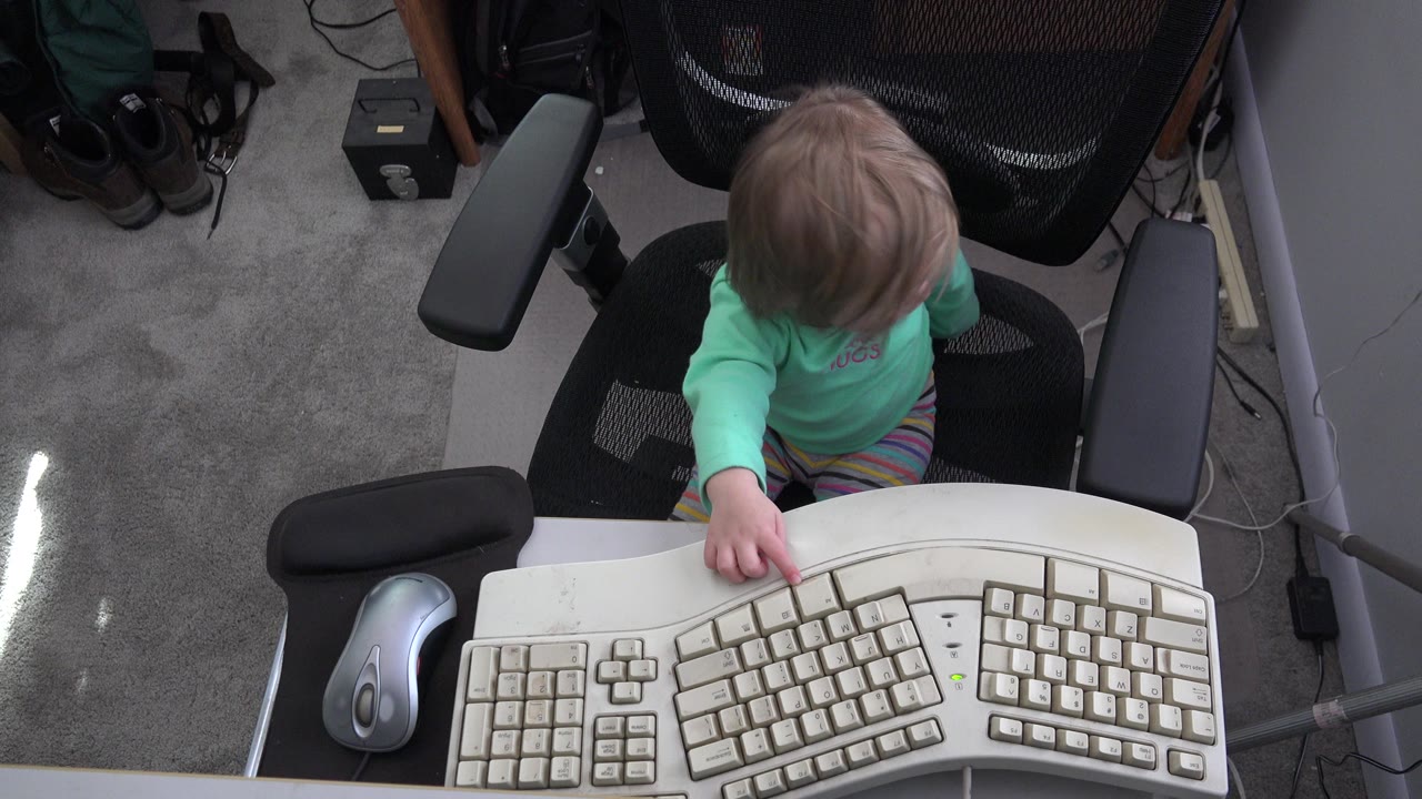 At 0:00 in Westminster, Colorado, a young girl sits below a desk edge and extends one hand toward a large beige keyboard, with a silver mouse and gray carpet nearby.