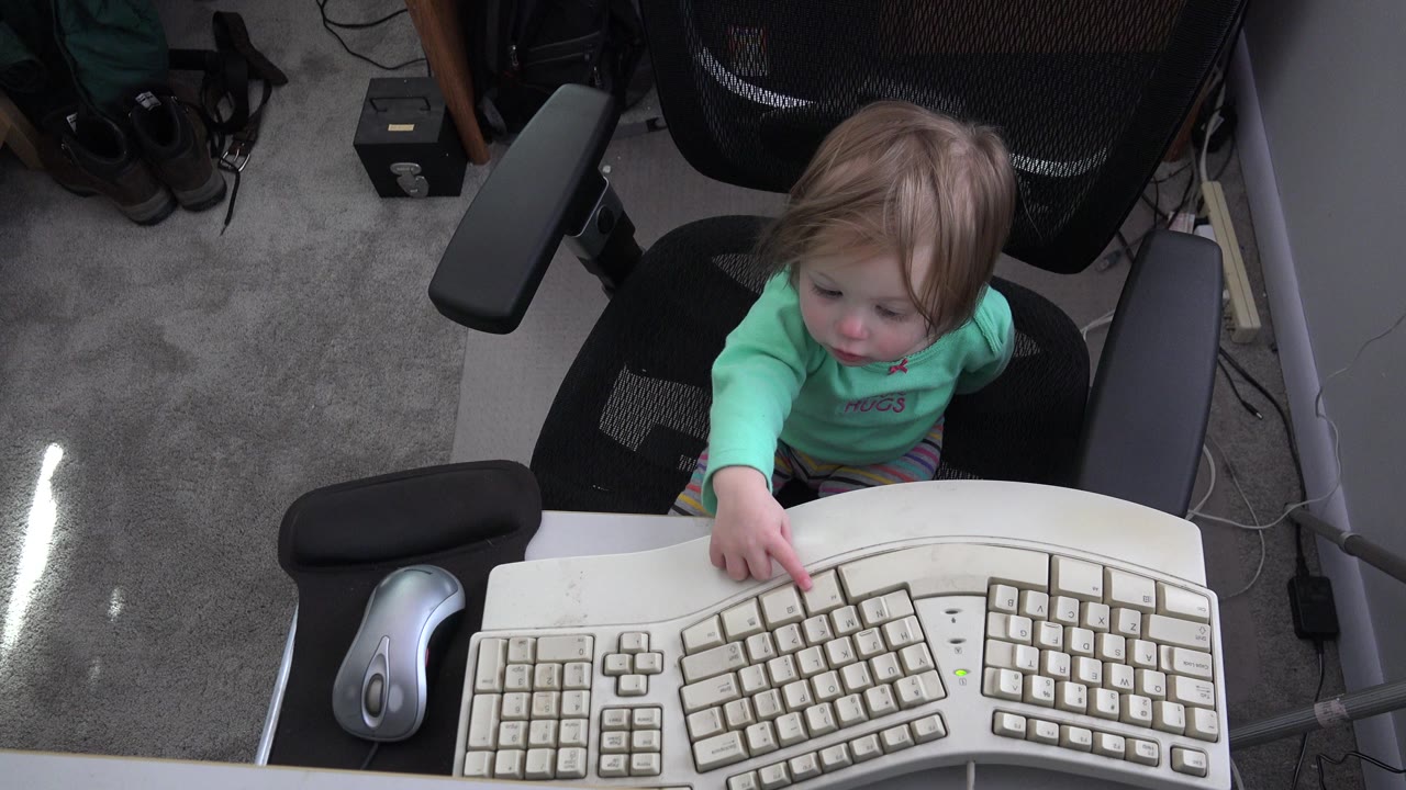 Toddler in an office chair reaching toward a white computer keyboard.