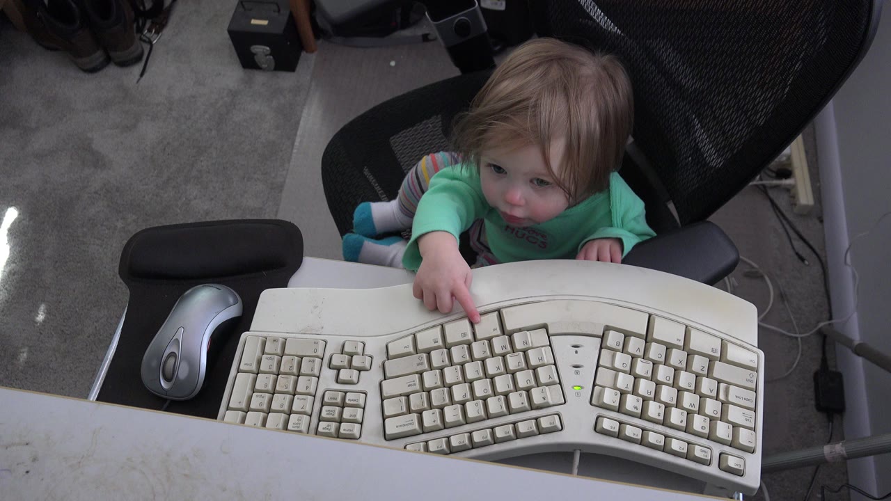 Young blonde girl in a chair reaching toward a white computer keyboard.