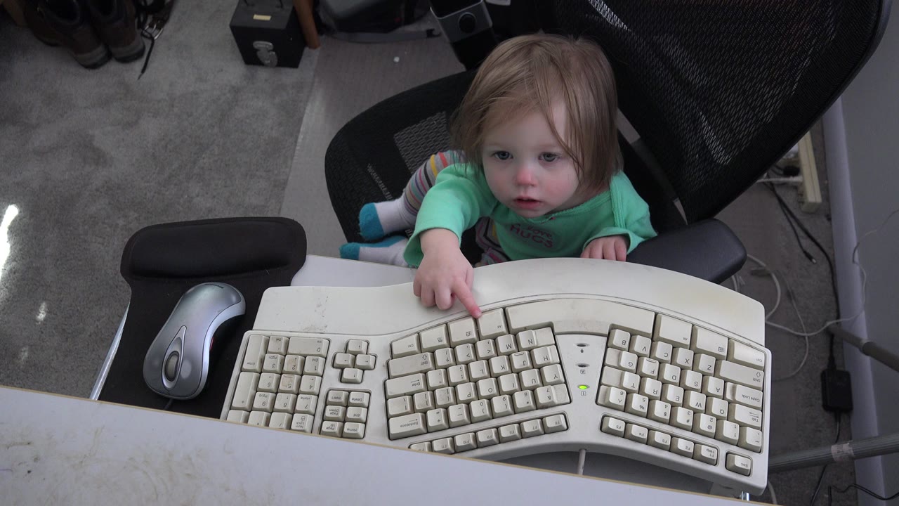 At 0:00 in Westminster, Colorado, a small child in a green shirt reaches from a black office chair toward a beige computer keyboard on a white desk.