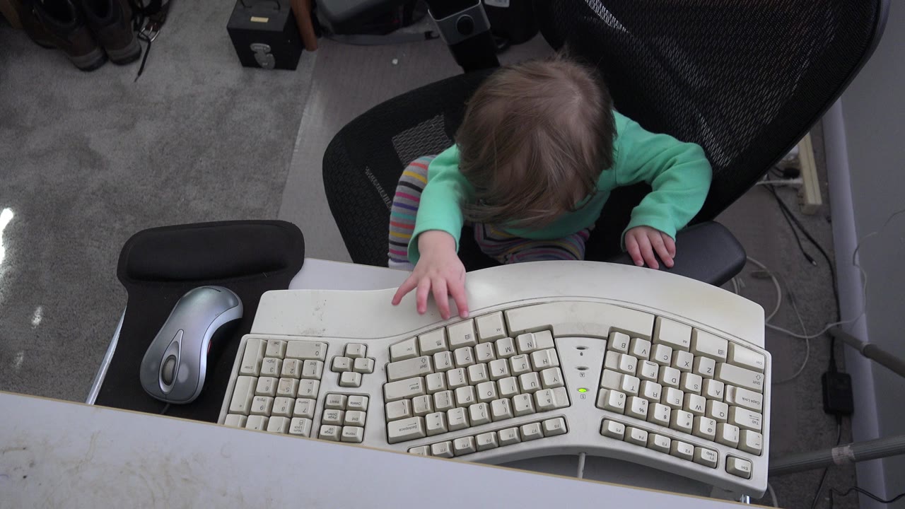 Toddler in an office chair reaching toward a computer keyboard.