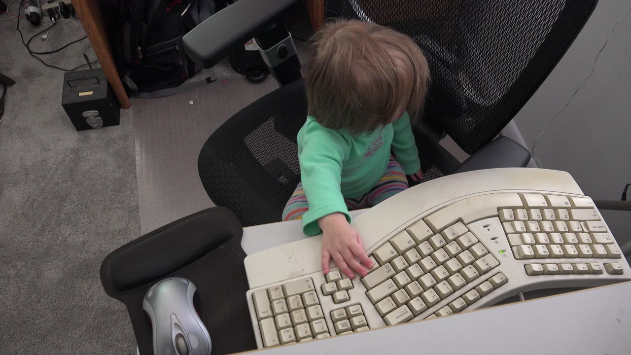 Young child reaches toward a beige computer keyboard from an office chair.