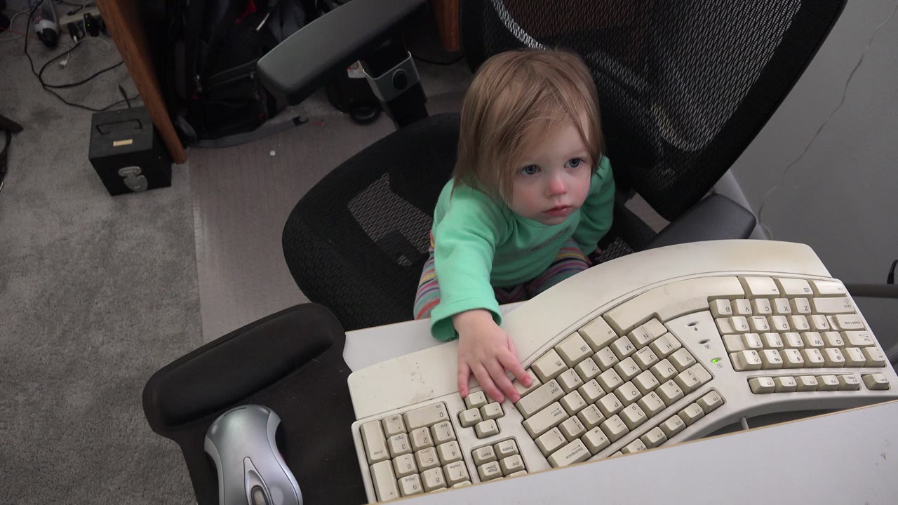 Baby girl reaches toward a beige computer keyboard from a black office chair.