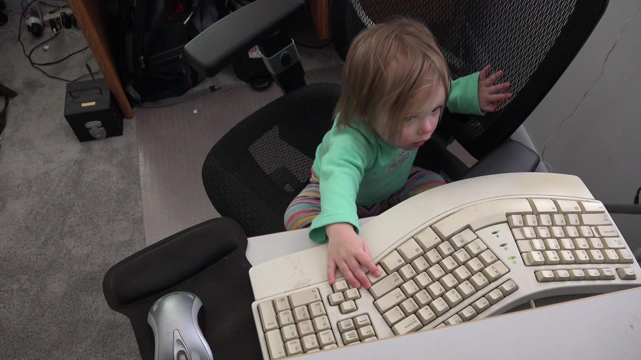 Toddler in an office chair reaching toward a computer keyboard on a desk.