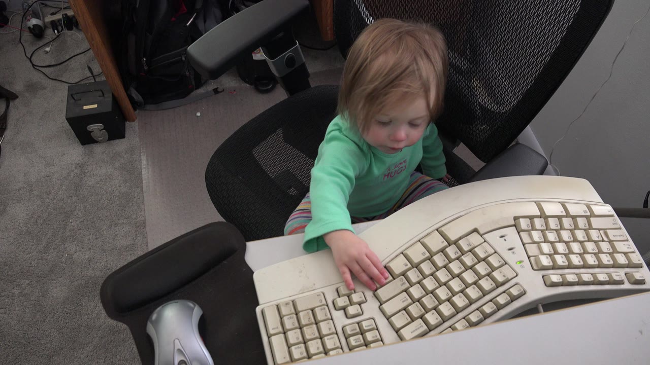At 0:01 in Westminster, Colorado, the child looks down while holding one hand near the split keys of the beige ergonomic keyboard.