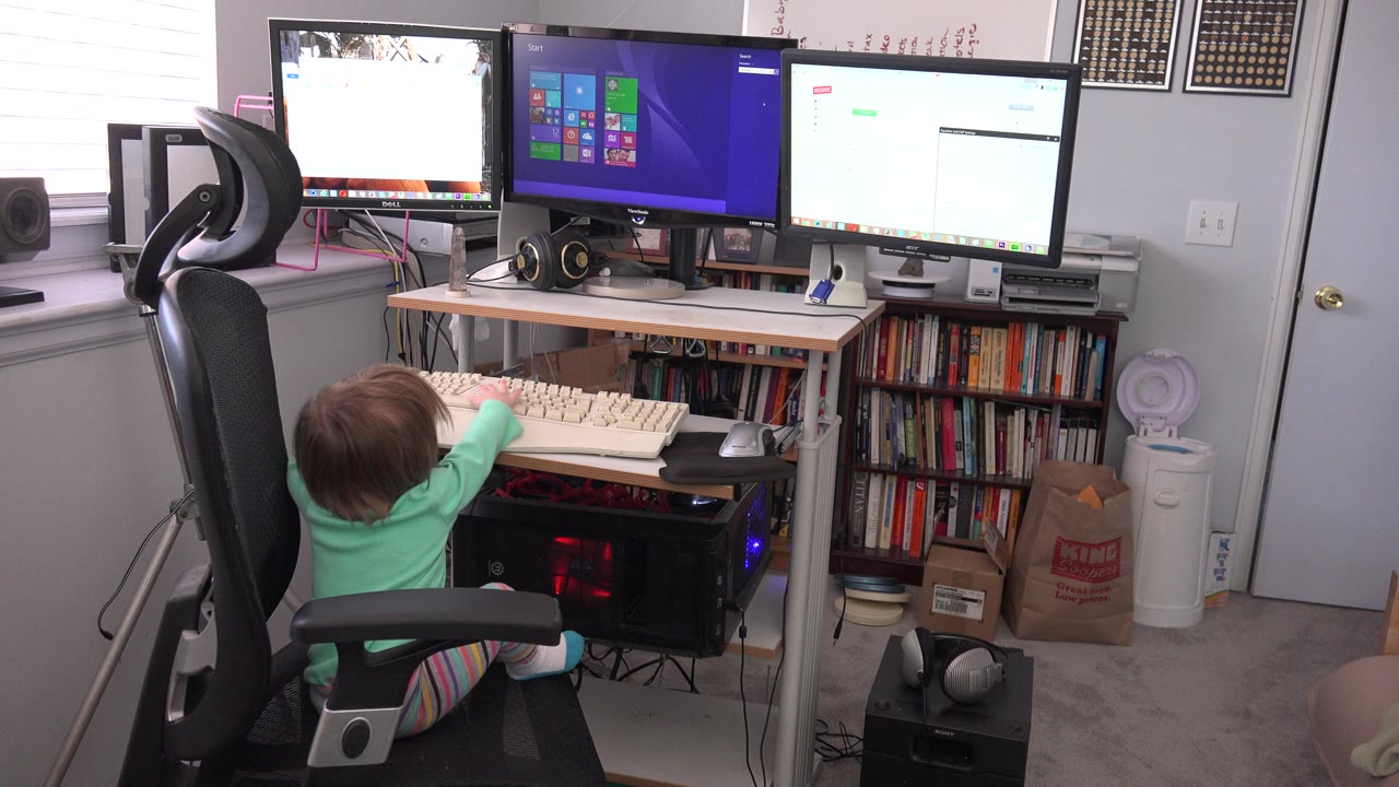 At 0:00, a toddler in a mint green shirt sits in a black office chair in a Westminster, Colorado home office, reaching toward a beige keyboard beneath three computer monitors.