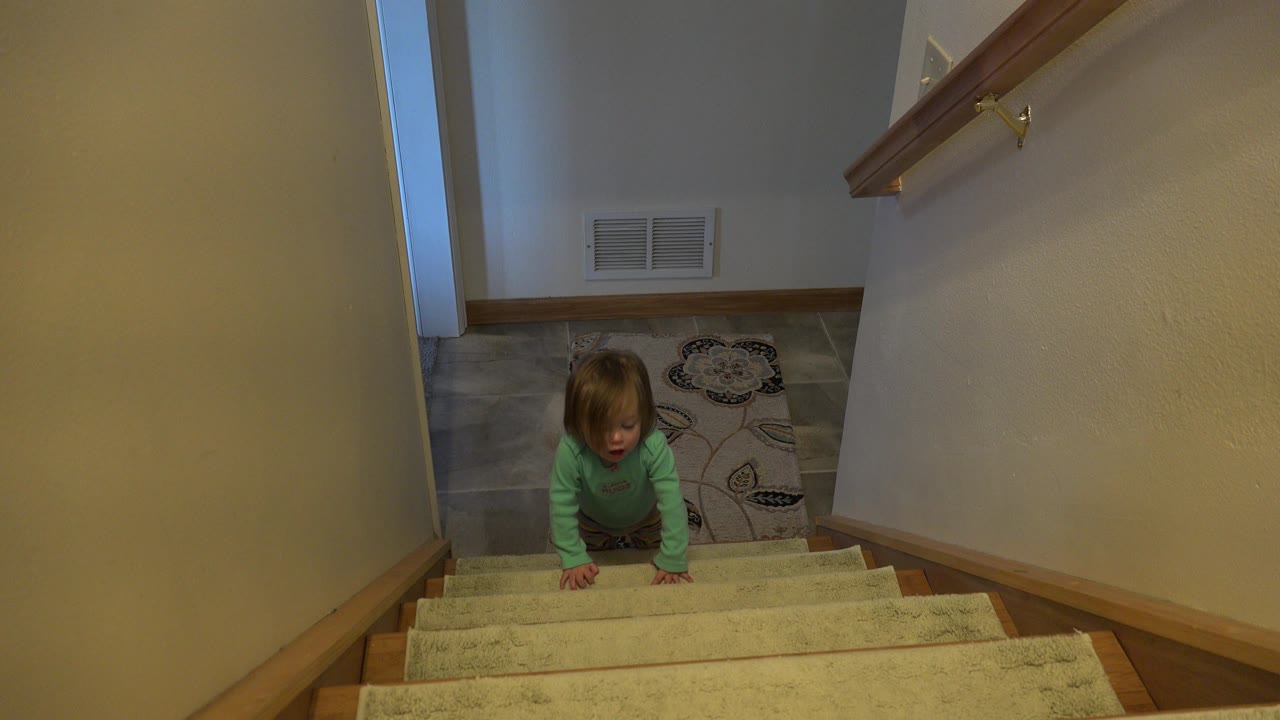 At 0:01, the toddler has moved slightly higher on the carpeted stairs, facing upward toward the camera while the hallway landing and patterned rug remain in the background.