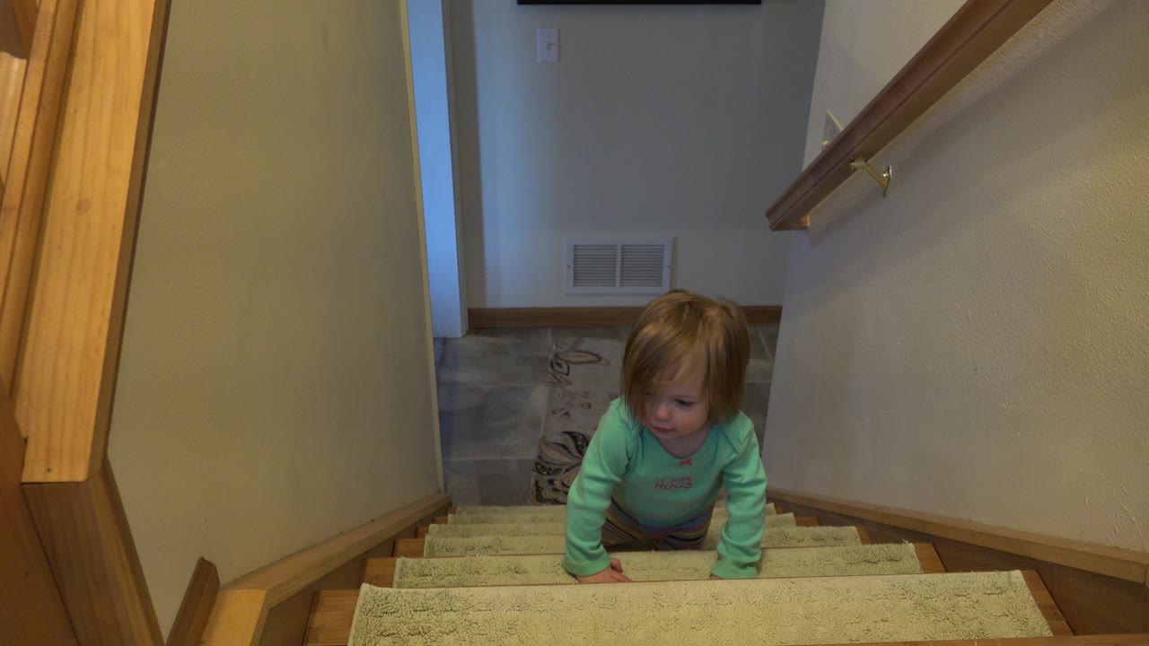 At 0:01 in Westminster, Colorado, the baby remains low on the carpeted steps, with the right-side handrail and beige stairwell walls framing the scene.