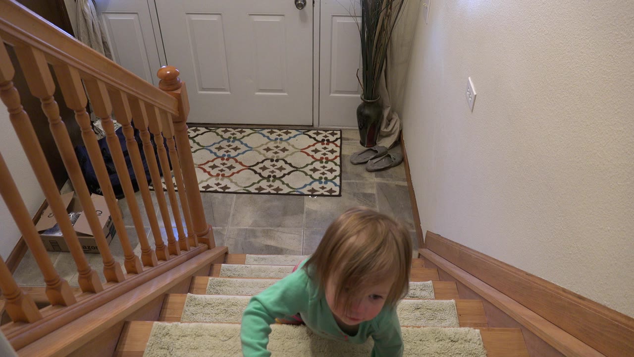 A young child in a green top appears at the lower edge of carpeted stairs inside a Westminster, Colorado home, with a wooden banister, tan wall, and white entry door visible.