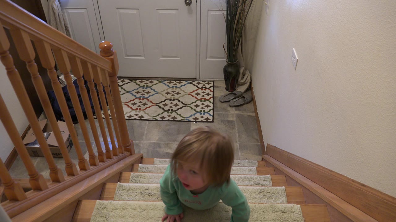 At 0:00 in Westminster, Colorado, a toddler in a mint green top is partway up carpeted interior stairs beside a wooden railing, with the front entry visible below.