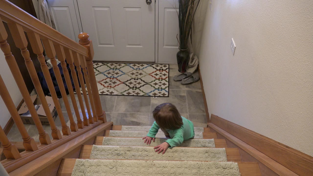 At 0:00, a young child in a green top climbs carpeted stairs inside a Westminster, Colorado home, with a wooden railing and entry door behind them.