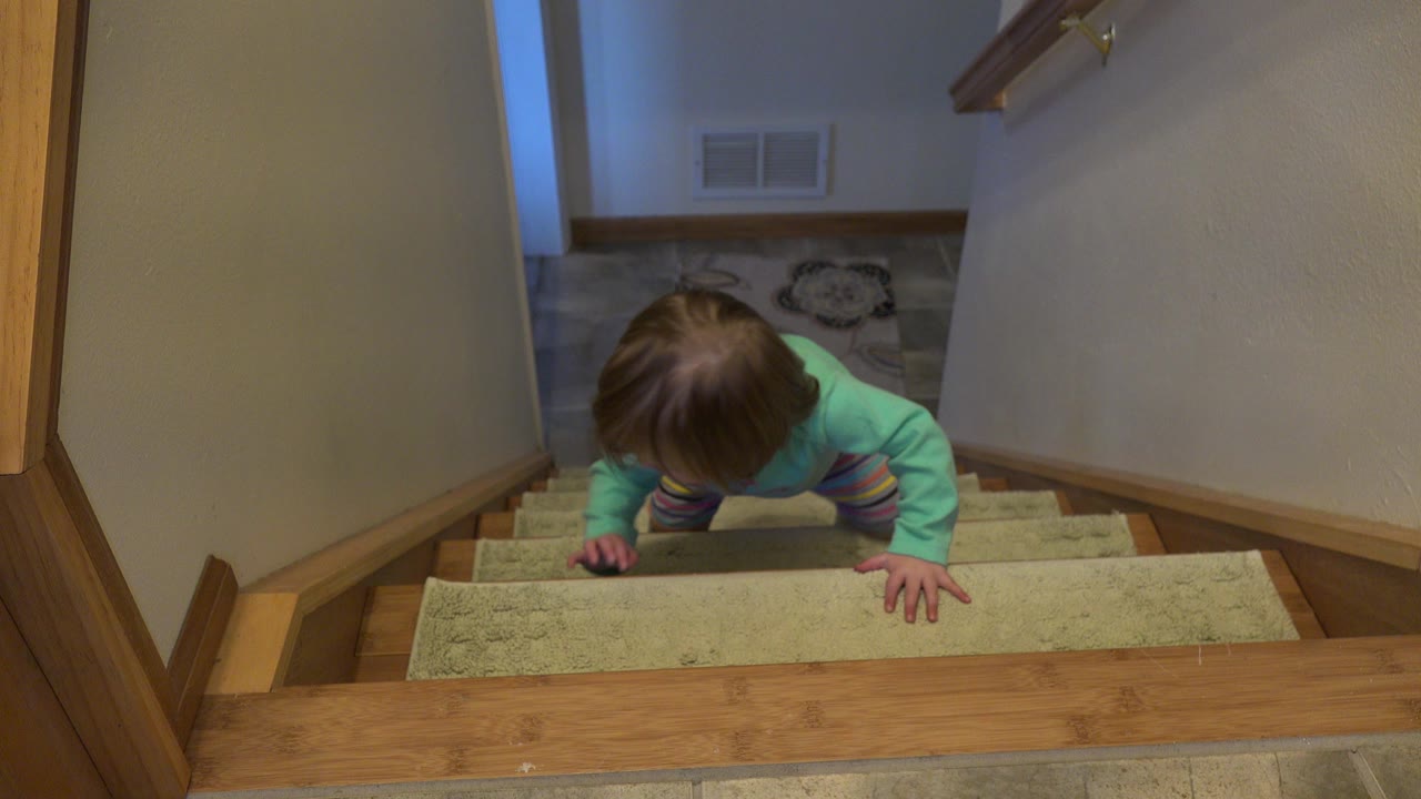 A small child in a turquoise shirt is near the top of beige carpeted stairs in a Westminster, Colorado home interior, with hands on the steps and wood trim around the landing.