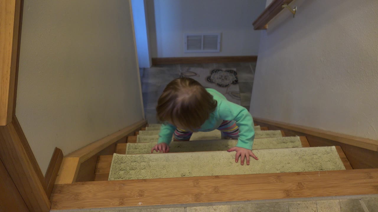 The child remains close to the upper landing on the carpeted staircase in Westminster, Colorado, with both hands braced on the steps and the lower hallway visible behind.