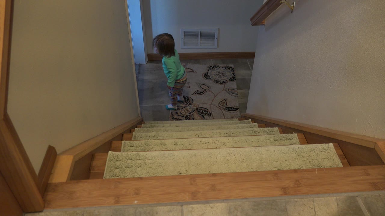 Young child standing on a landing below carpeted stairs.