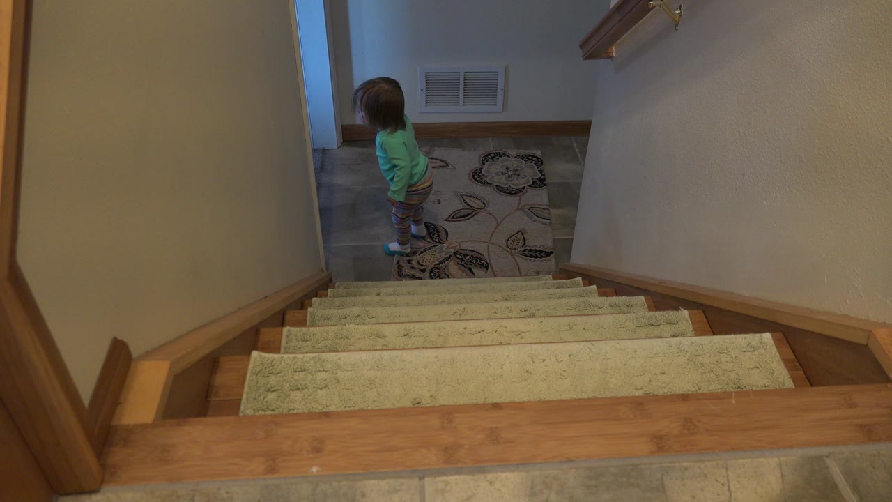 At 0:01 in Westminster, Colorado, the child remains on the tiled landing below the stairs, turned slightly toward the adjacent doorway.