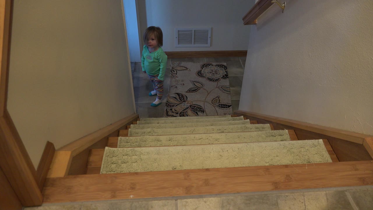 The child remains at the lower landing, facing toward the stairs with the carpeted steps filling the foreground inside the Westminster home.