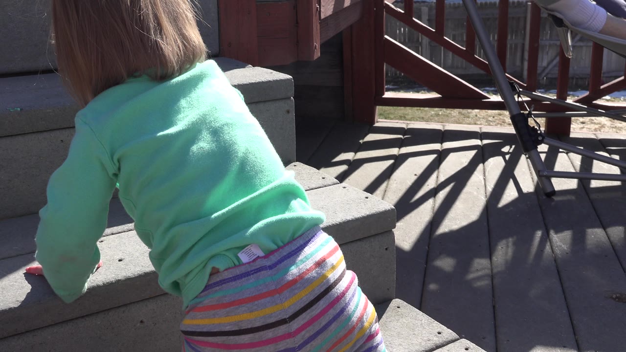 At 0:01, the toddler leans farther onto the step, with red railing posts, dark diagonal shadows, and a concrete stair edge visible around the child.