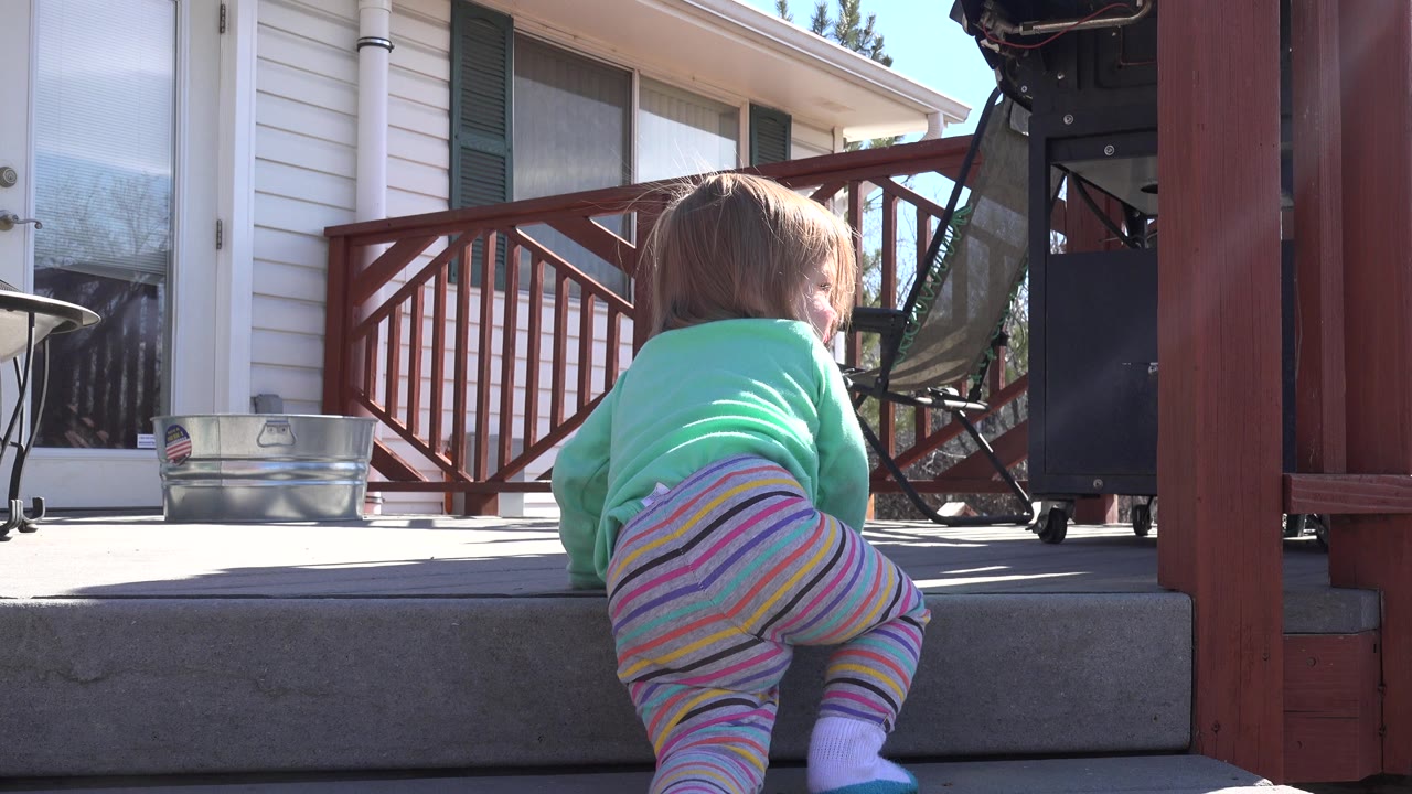 Child crawling toward a concrete patio step beside a red deck railing.