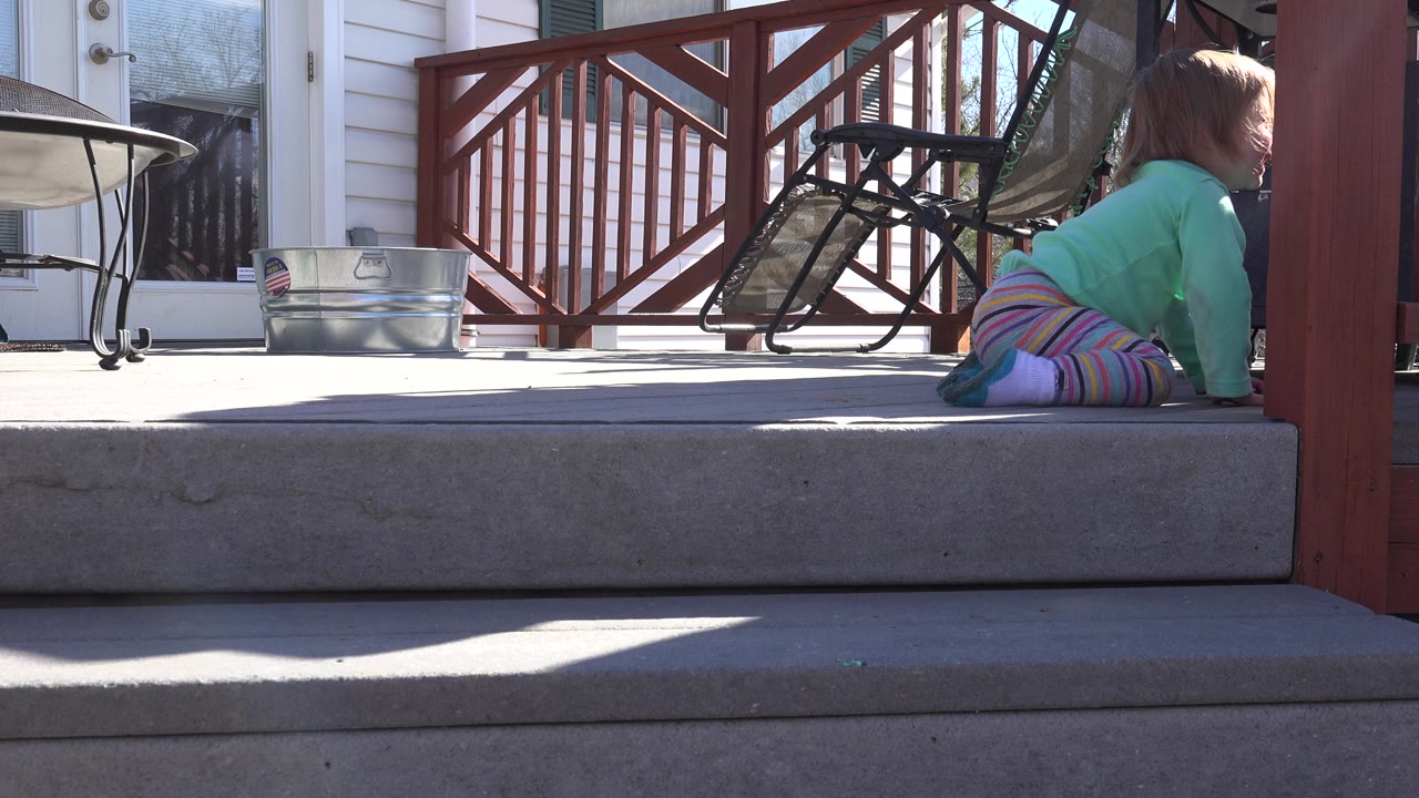 A young child in a green top and striped pants is low to the terrace floor in Westminster, Colorado, with concrete steps in front and red porch railing behind.