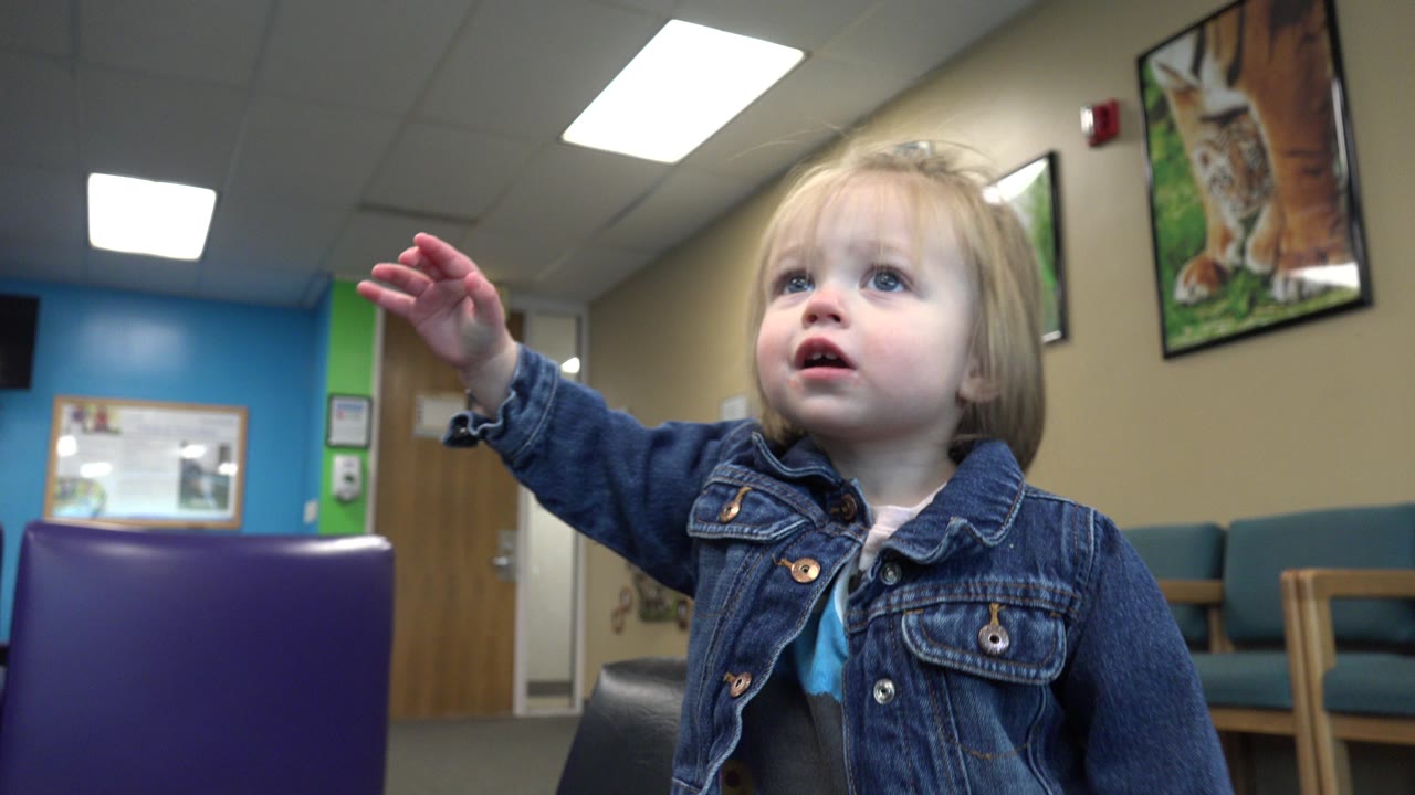 A blonde toddler in a denim jacket raises her left hand while looking upward in an indoor Denver waiting area with purple and teal chairs behind her.