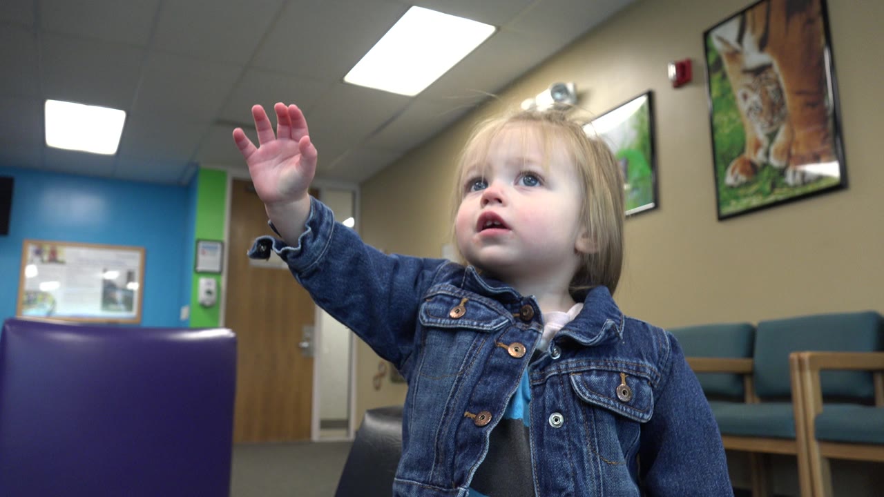 A blonde child in a denim jacket lifts her hand higher, with waiting-room chairs, animal posters, and bright ceiling lights visible in the Denver interior.
