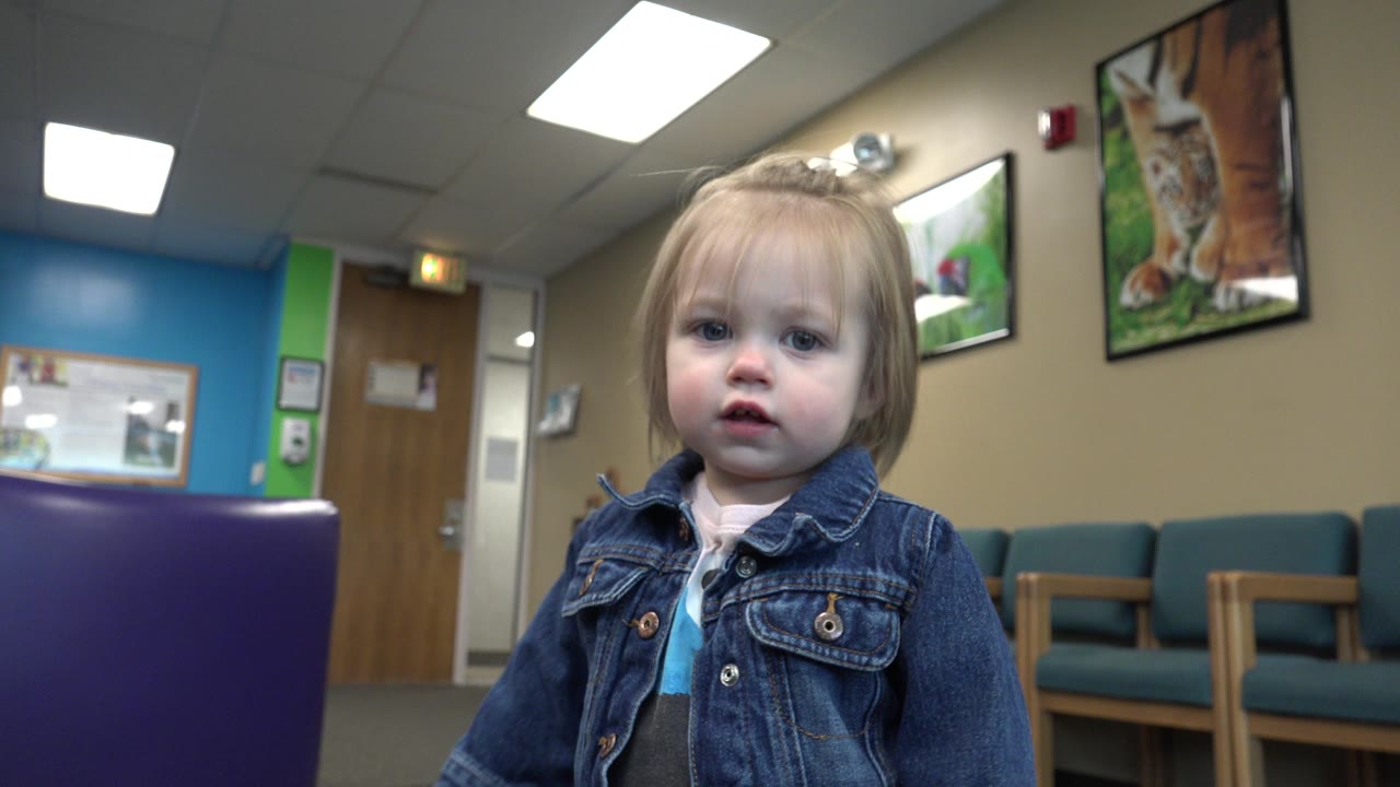 The child appears slightly farther right in the frame inside the Denver doctor's office waiting room, with fluorescent lights overhead and framed artwork on the tan wall.