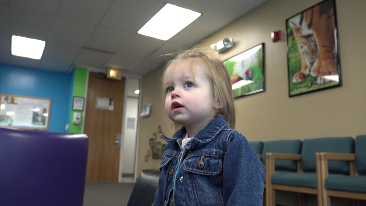 The child remains near the foreground chair in the Denver interior, looking slightly up while teal chairs and wall-mounted framed pictures fill the background.