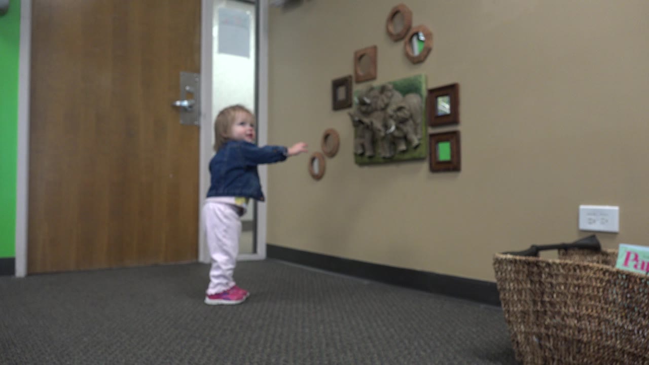 In Denver, Colorado, the child remains near the door, smiling and pointing across the carpet toward the small framed pieces and raised plaque on the wall.