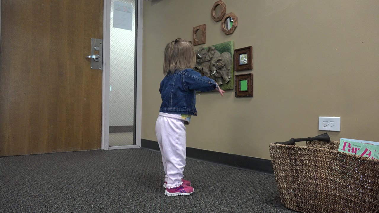 Toddler in denim jacket facing wall decor in a carpeted room.