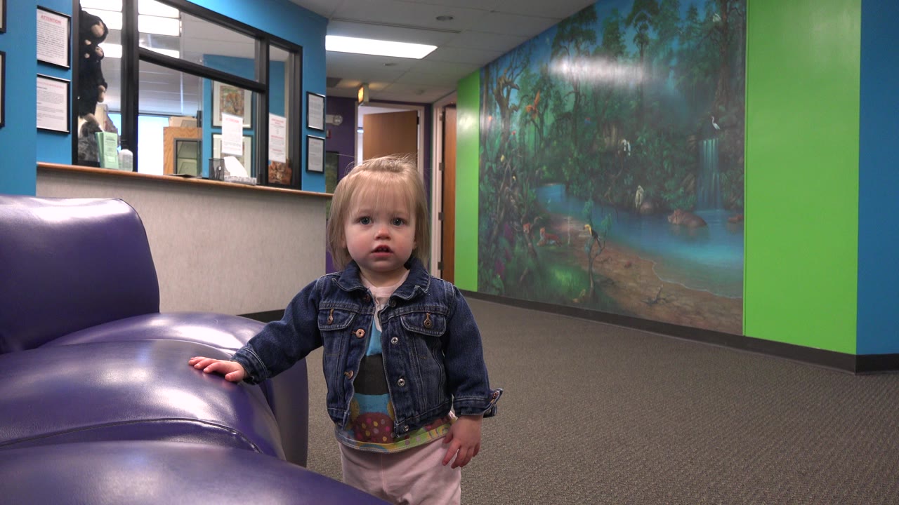 A blonde toddler in a denim jacket stands near the left end of a purple sofa, facing the camera with blue and green walls and a mural behind her.