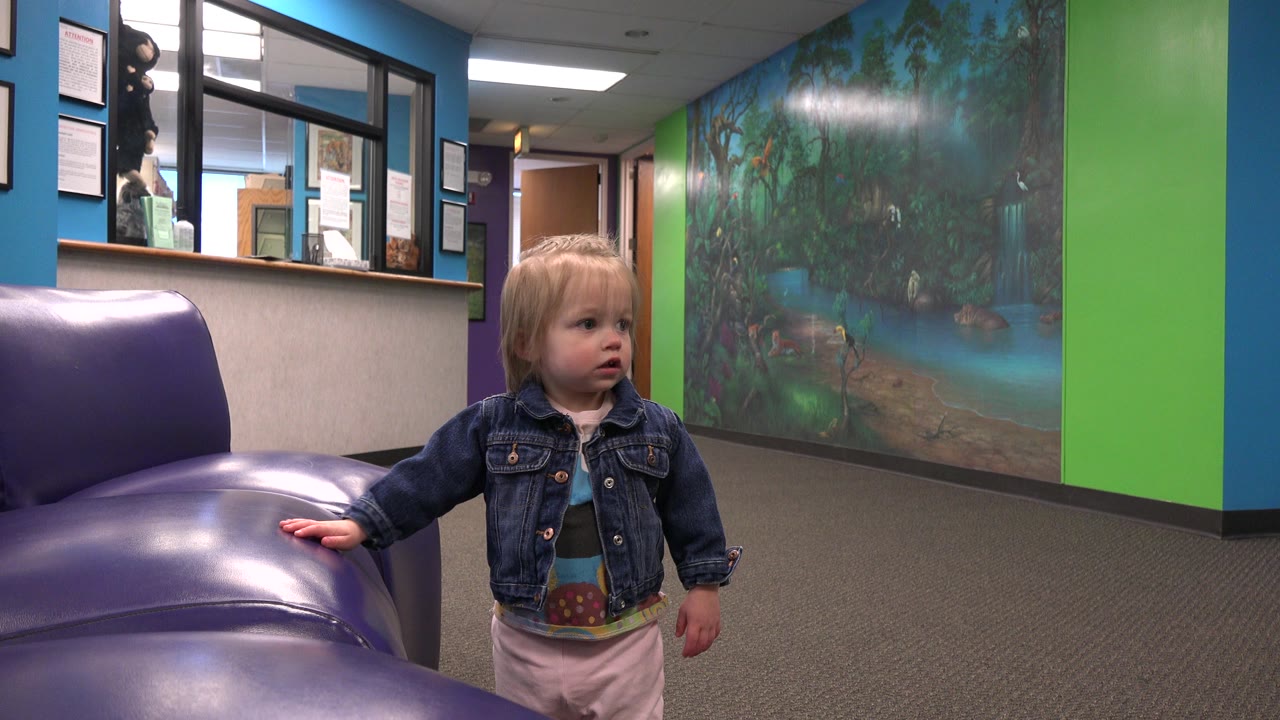 The toddler turns her head to the right while keeping one hand on the purple sofa, framed against the green column and painted mural wall.