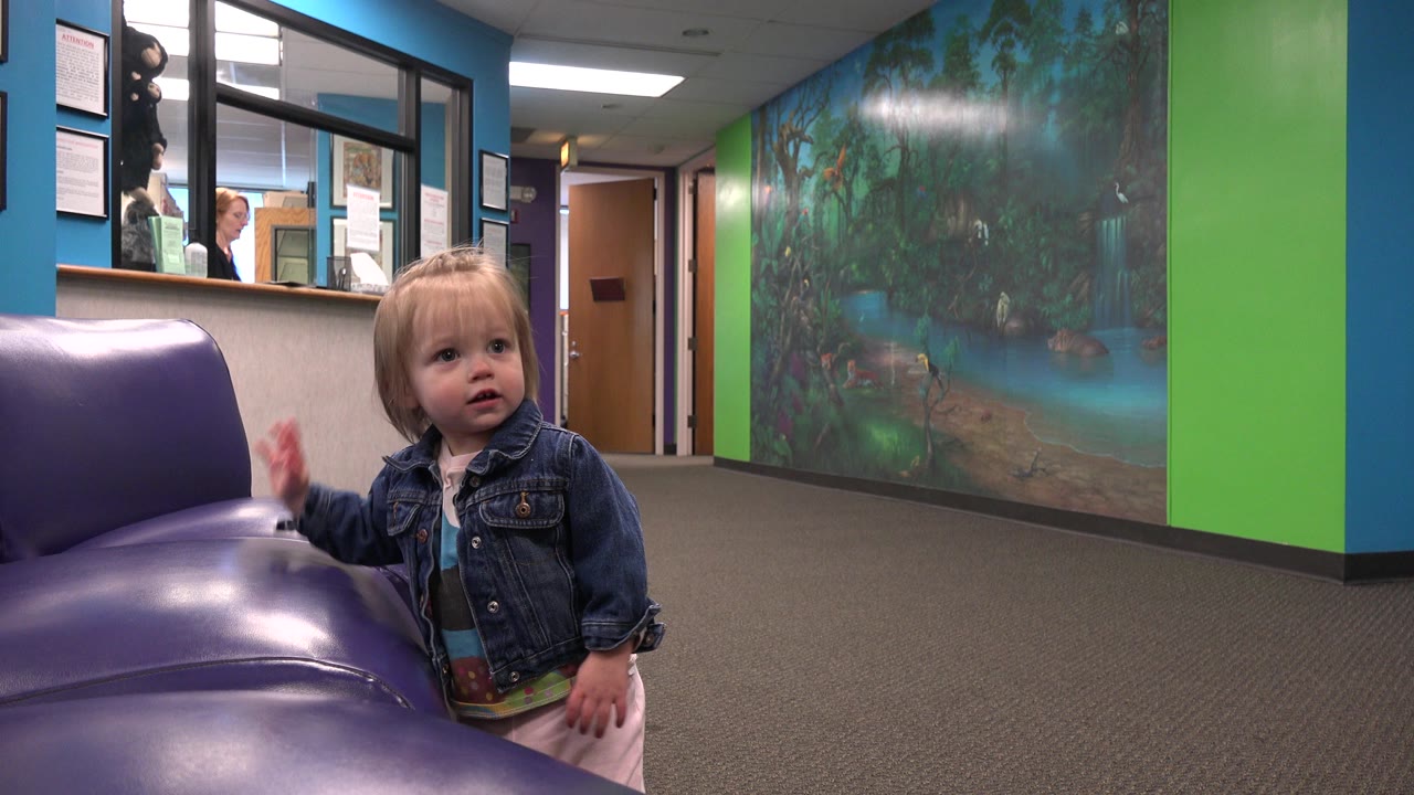 A blonde toddler in a denim jacket stands near purple vinyl chairs in a colorful waiting room, with office windows and a wall mural behind her.