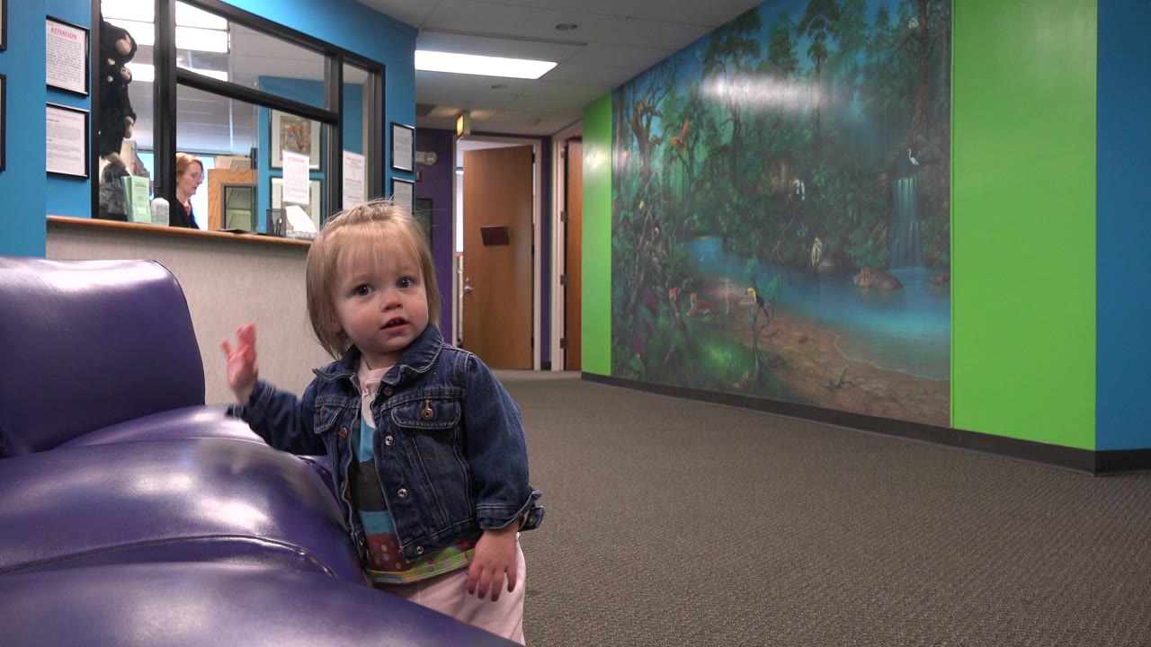 Toddler raises one hand near a purple chair.