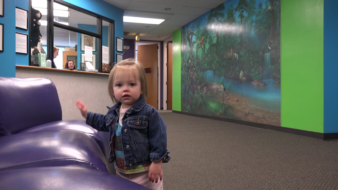 The child stands close to the purple chair and looks forward, with reception-style interior windows, overhead fluorescent light, and bright wall colors behind her.
