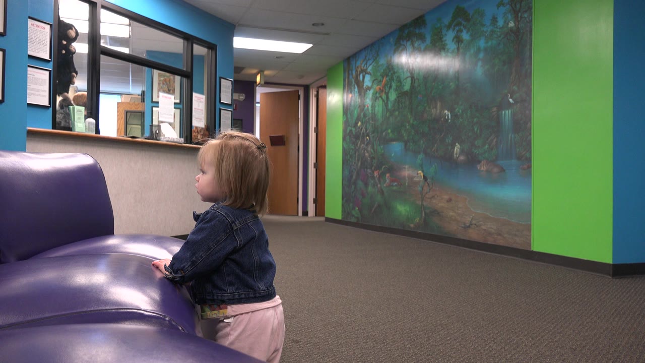 Blonde toddler beside a purple bench in a colorful doctor waiting room.