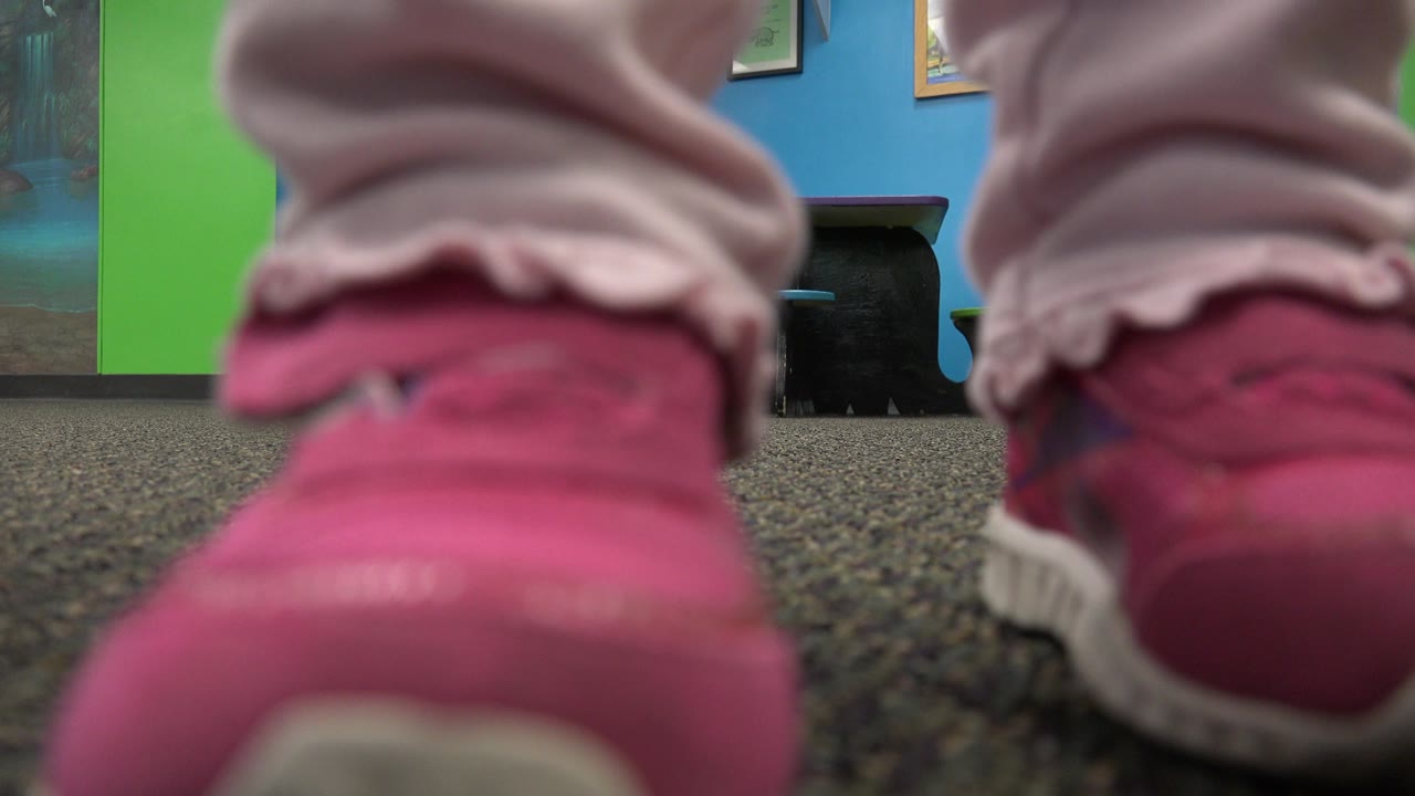 Floor-level close-up of small pink trainers and pale pants standing on gray carpet, with a green wall, blue wall, dark furniture, and framed pictures blurred in the background.