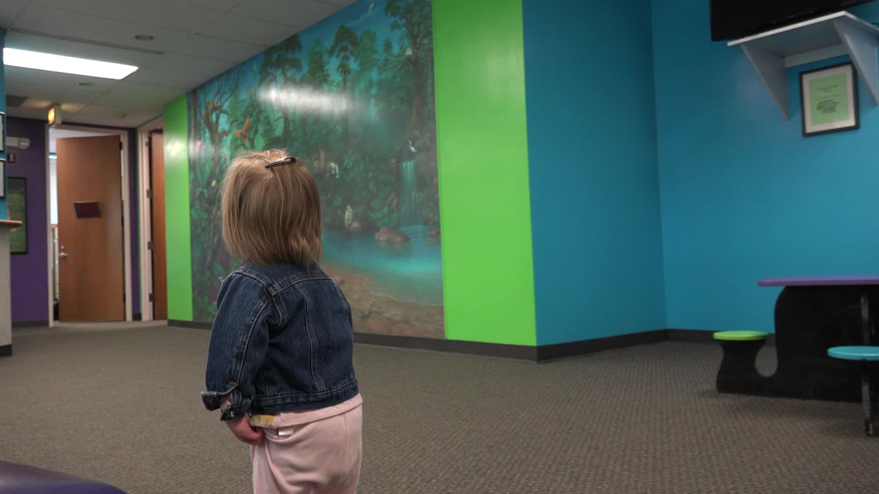 A young child in a denim jacket stands with their back to the camera in a bright waiting room with teal and green walls, a large mural, carpet, doors, and purple seating at the edges.