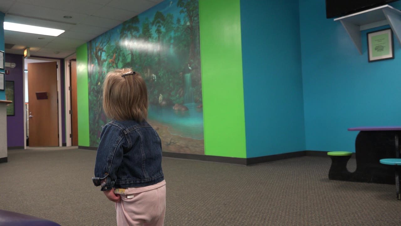 The child remains turned away near the center of the frame, with the teal wall, green vertical stripe, large mural, closed doors, carpet, and low seating visible around the waiting room.