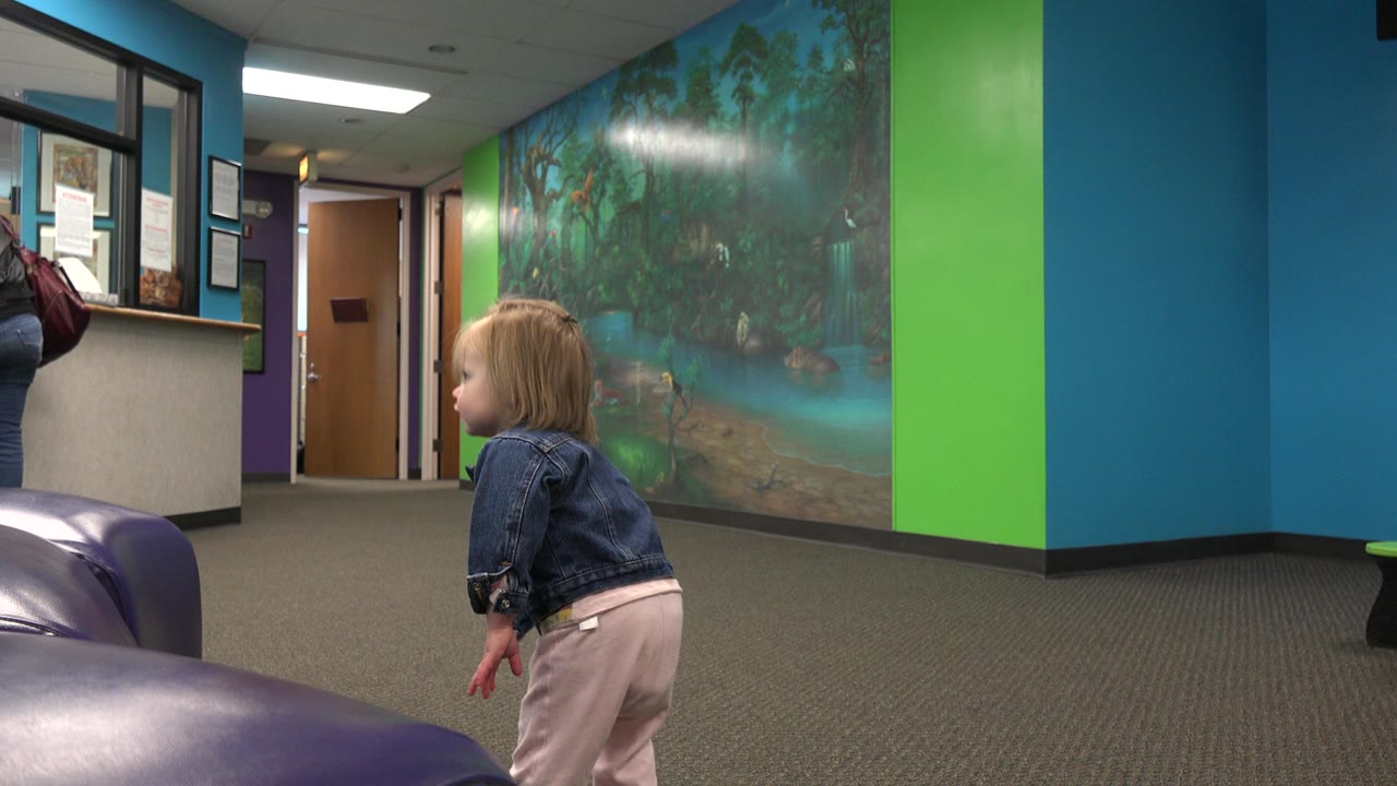 A blond toddler in a denim jacket stands near purple padded seating, facing left in a teal and green indoor hallway with a wall mural behind her.