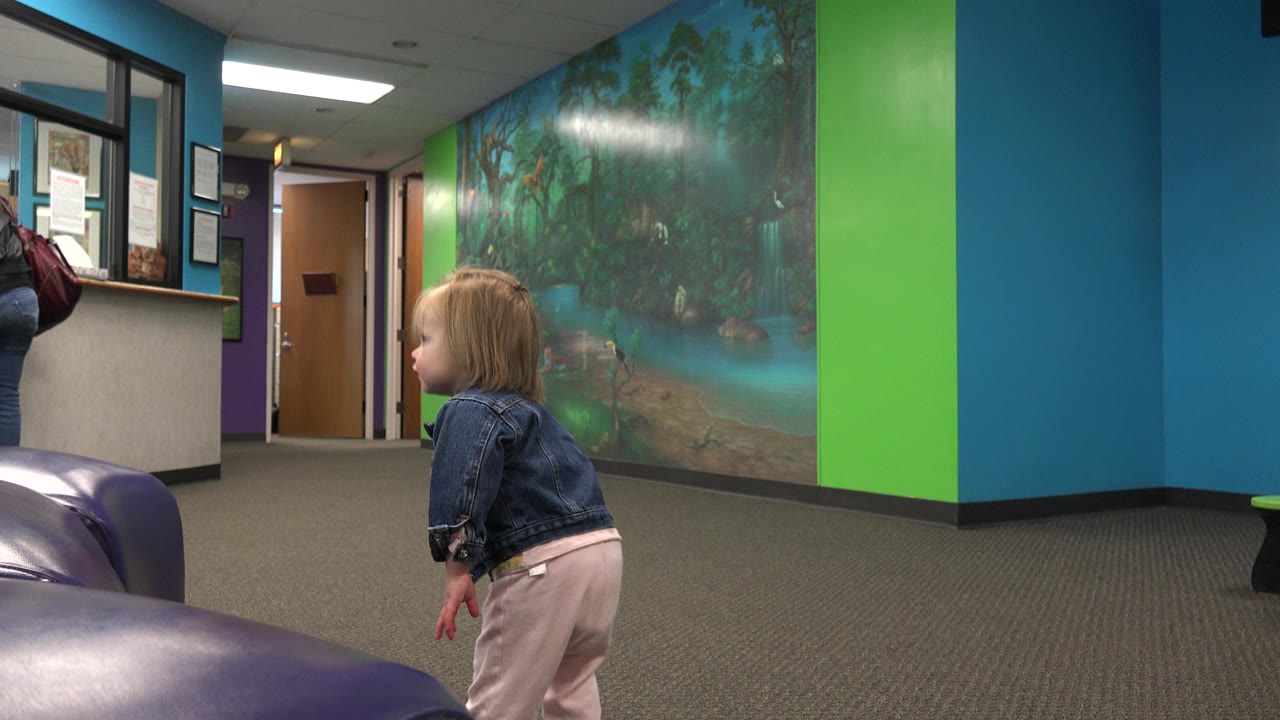 Toddler stands near purple seating with mural wall and doors behind her.