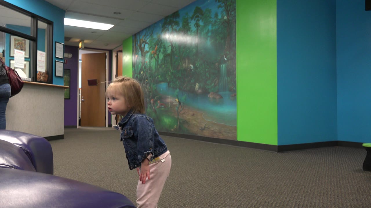 The toddler turns her face more toward the camera while standing in the foreground, with the teal wall, green panel, mural, carpet, and open hallway door behind her.