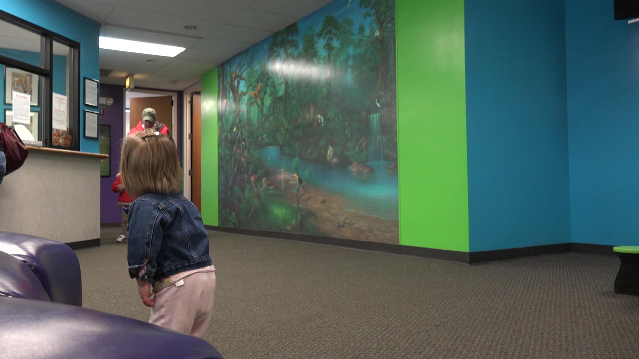 Toddler in a denim jacket stands indoors facing an adult near a doorway.