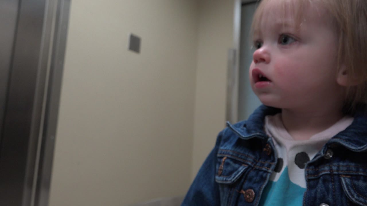 Blond toddler in a denim jacket looking left in a hallway.