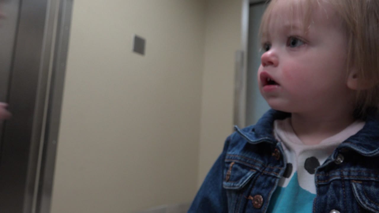 The toddler remains near the hallway wall in Denver, Colorado, with their face turned left and the corridor surface filling the background.