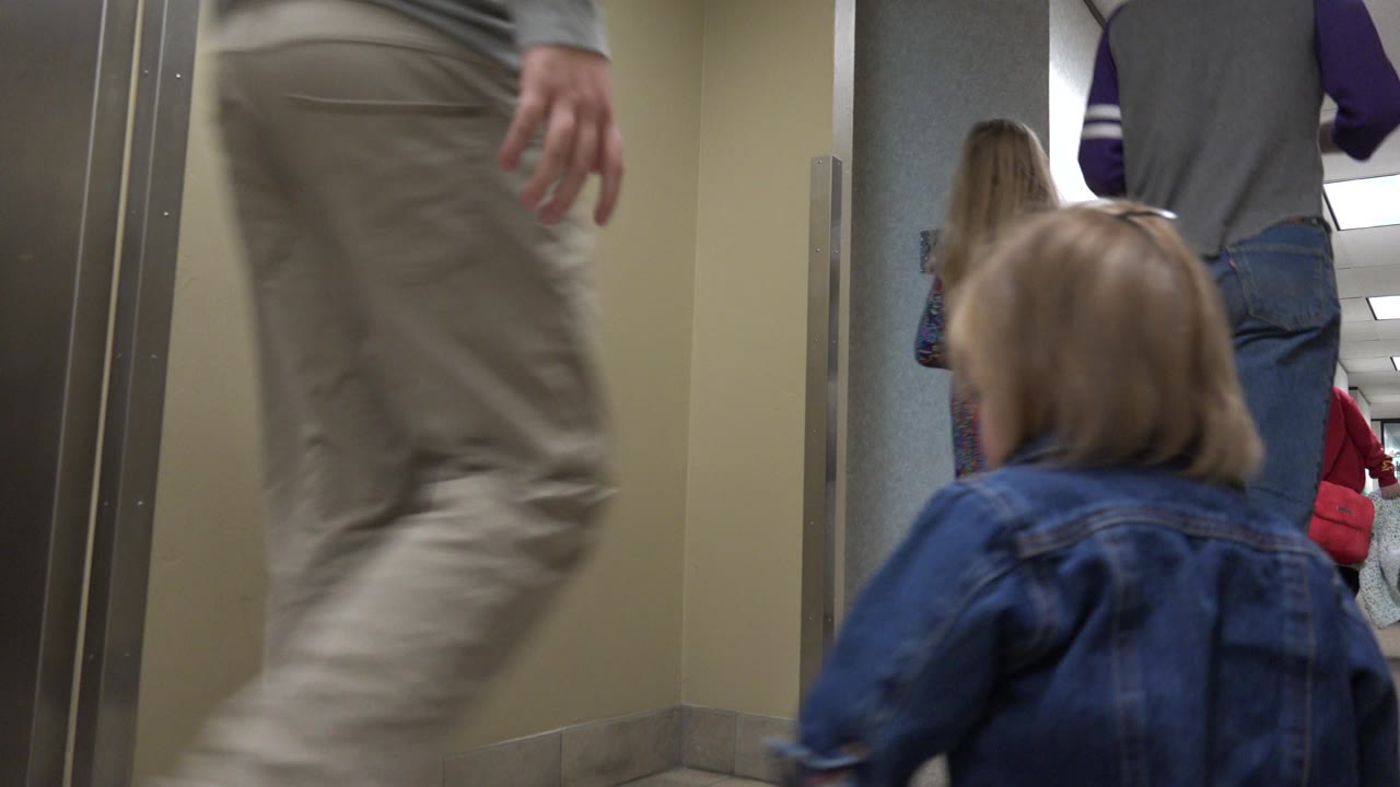 The toddler is seen from behind in the Denver corridor, wearing a blue denim jacket while adults walk ahead under fluorescent ceiling lights.