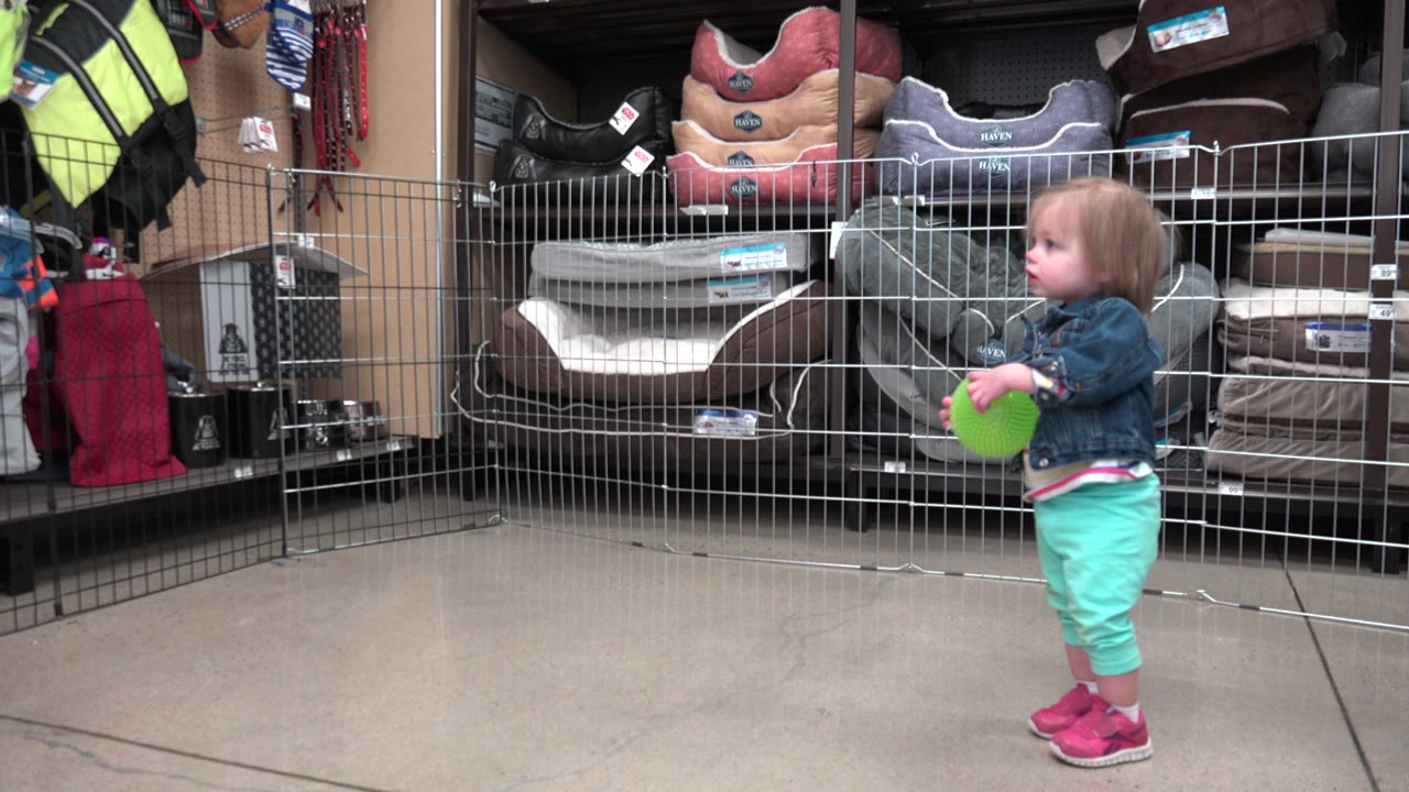 Toddler holding a green ball beside wire railings in a store aisle.