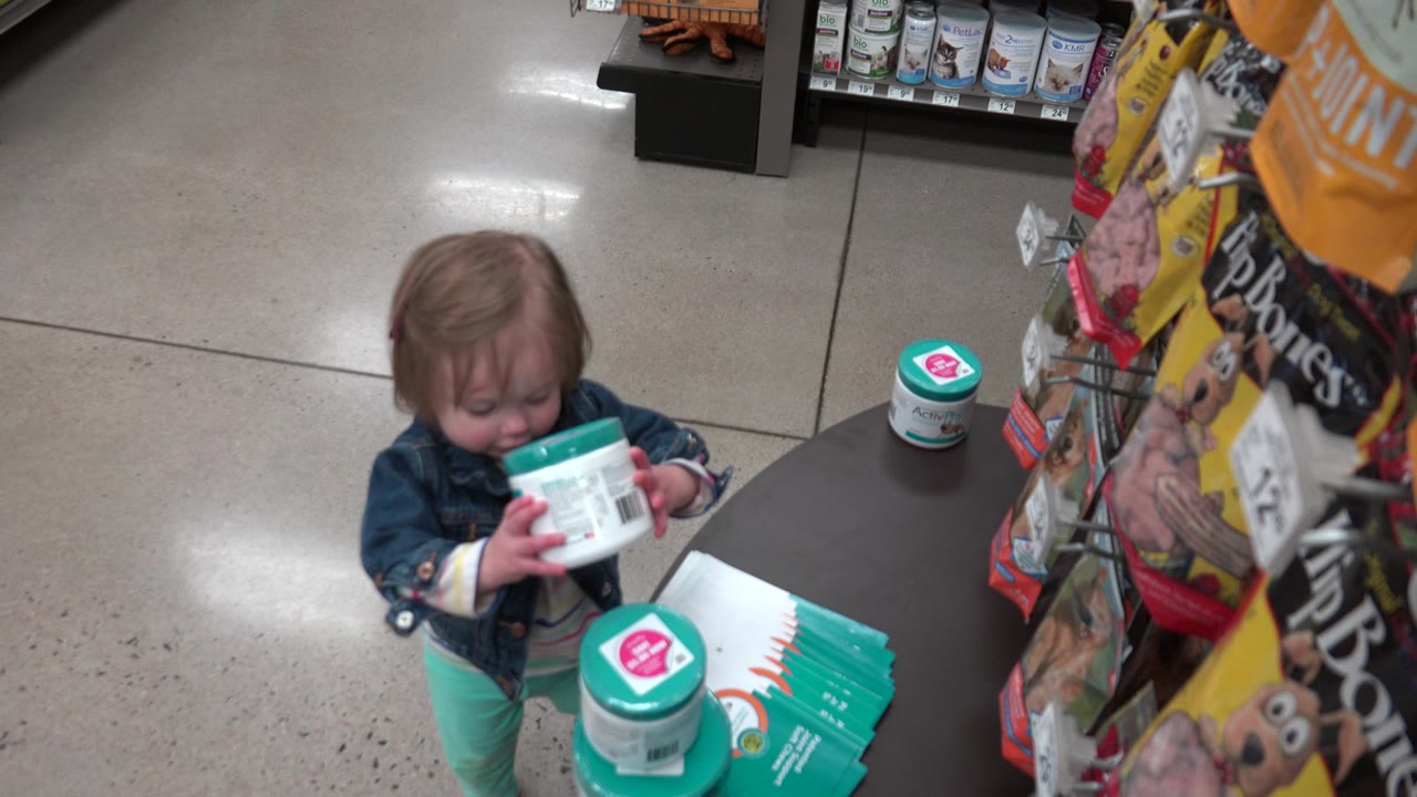 The child looks down while holding a green-lidded white container, with another container on the display and shelves of products visible behind her in the Denver retail setting.