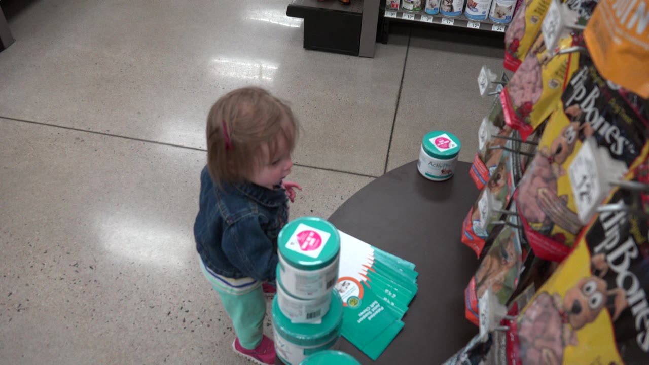 A young child in a denim jacket stands beside a black display table with teal and white packages in a Denver grocery store aisle.