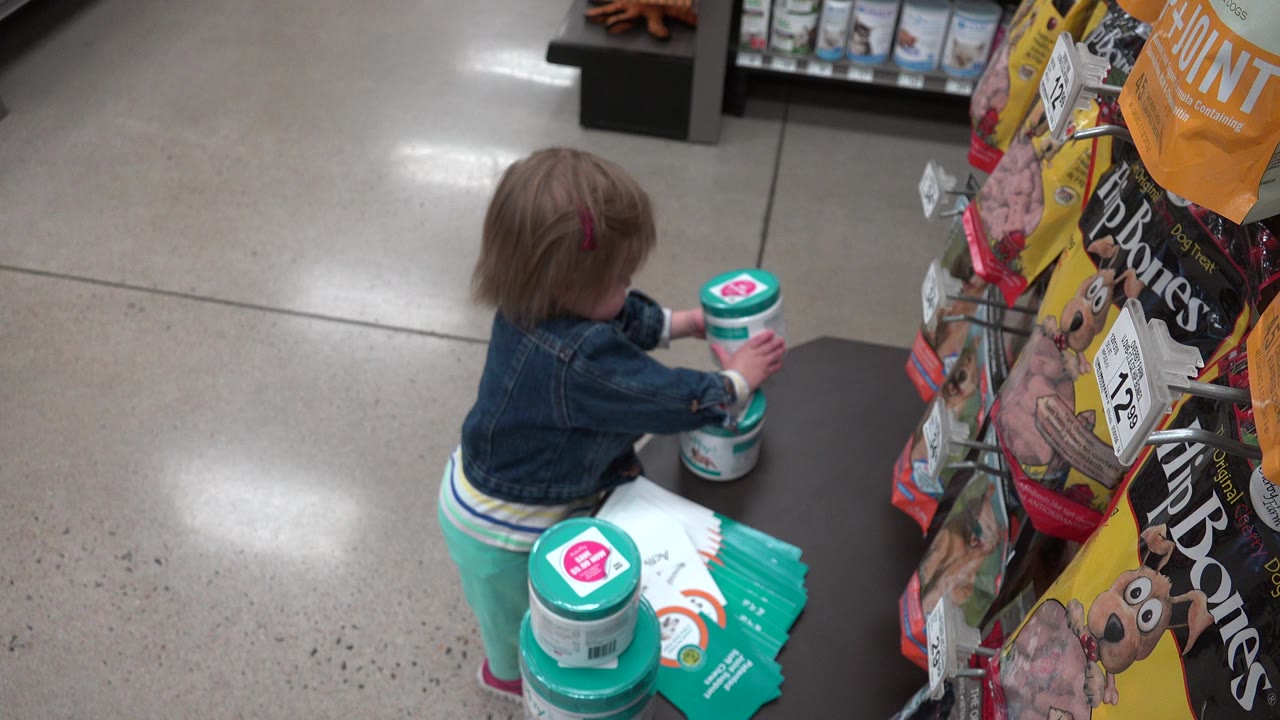 In a Denver supermarket aisle, a small child in a denim jacket reaches toward a teal-lidded container beside a display of packaged goods.